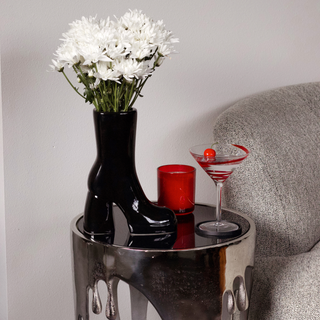A black boot vase filled with white flowers sits on a table next to a martini glass on a table. The table is positioned in front of a couch.
