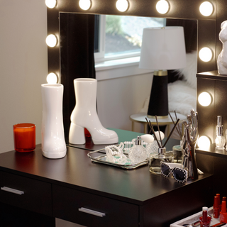 A vanity with a rectangular mirror with bright lights. There is a white boot vase sitting on the table next to the mirror.