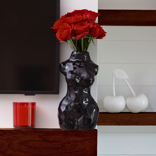 A black glossy ceramic vase shaped like a woman’s torso sits on a shelf. The vase is filled with red roses