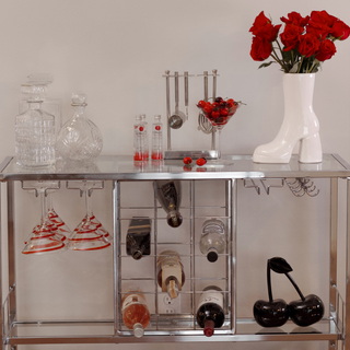 A bar cart with a white ceramic boot vase filled with red flowers and a martini glass with cherries inside. The bar cart also has several wine bottles and martini glasses stored on it.
