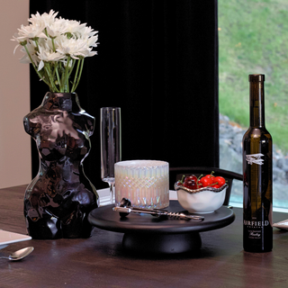 A glossy black ceramic vase shaped like a woman's torso sits on a dining table next to a white candle, a bowl of red cherries, wine opener, and a bottle of wine. The vase is filled with a bouquet of white flowers.