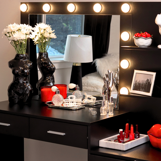 A lighted vanity table with a mirror. There are white flowers in a glossy black body-shaped vase and perfume bottles, a candle, and nail polish on the vanity table.