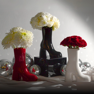 Three ceramic vases shaped like high-heeled boots sit on a table. From left to right: red boot with white flowers, black boot with white flowers, white boot with red flowers. The vases are displayed next to shiny disco balls.