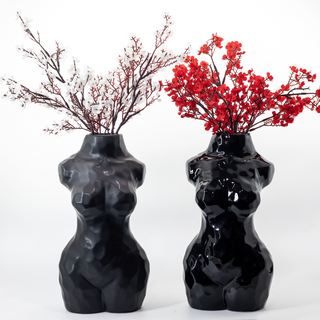 Two black, ceramic vases shaped like women's torsos. The vase on the left has a matte finish and is holding white flowers in it, while the vase on the right has a glossy finish and is holding red flowers.