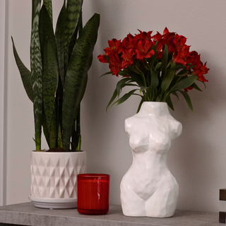 A glossy white, ceramic vase shaped like a woman’s torso holding a bouquet of red flowers. The vase sits on a table next to a potted plant and a candle.