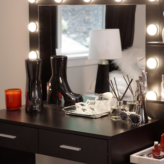 A vanity table with a rectangular mirror with bright lights. There is a black ceramic boot vase sitting on the table next to the mirror.