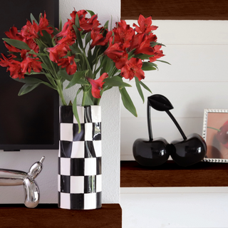 A black and white checkered vase. The vase has a wide opening and is filled with a bouquet of red roses. The vase sits on a light colored wooden table. To the right of the table is a picture of a black cherry.