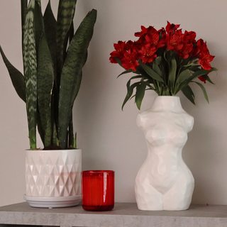 A matte white, ceramic vase shaped like a woman’s torso holding a bouquet of red flowers. The vase sits on a table next to a potted plant and a candle.