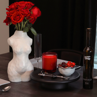 A matte white vase shaped like a female body sitting on a dining room table holding red roses. To the right of the vase is a candle, wine opener, a bowl of red cherries, and a bottle of wine.