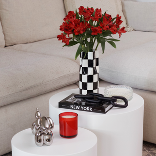 A close-up photo of a black and white checkered ceramic vase. The vase has a wide opening and is filled with a bouquet of red roses with green thorns. The vase sits on a white table with a book that reads "New York".