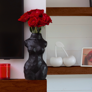 A black matte ceramic vase shaped like a woman’s torso sits on a shelf. The vase is filled with red roses.