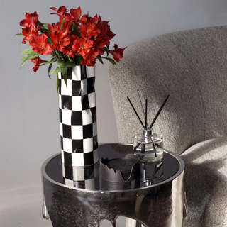 A black and white checkered ceramic vase with a wide opening sits on a light colored table. The vase is filled with a bouquet of red roses with green thorns.