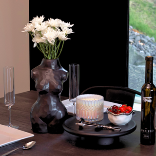 A matte black ceramic vase shaped like a woman's torso sits on a dining table next to a white candle, a bowl of red cherries, wine opener, and a bottle of wine. The vase is filled with a  bouquet of white flowers.