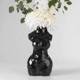 A black ceramic vase in the shape of a woman’s torso. The vase has a smooth, glossy finish and is filled with a bouquet of white flowers