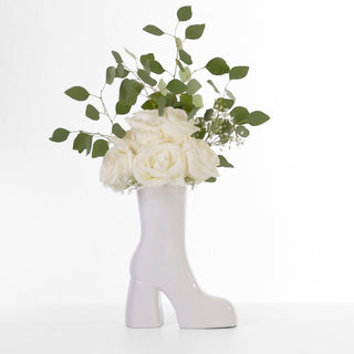 A white ceramic boot vase in the shape of a high heel. The vase is filled with a bouquet of white roses.