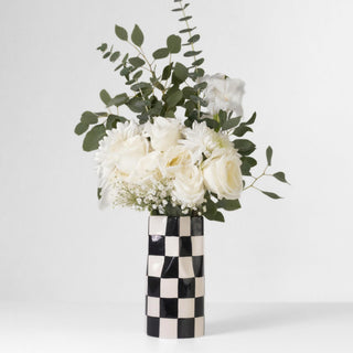 A black and white checkered vase with a wide opening sits on a surface. The vase holds a bouquet of white flowers.