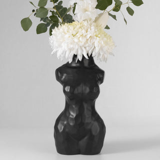 A black ceramic vase in the shape of a woman’s torso. The vase has a smooth, matte finish and is filled with a bouquet of white flowers
