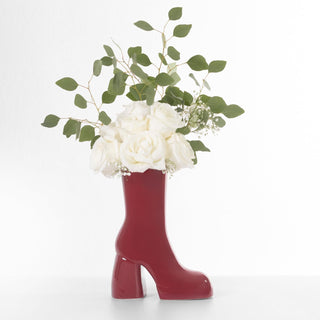 A red boot-shaped vase made of eucalyptus cinerea sits on a white background. The vase is filled with a bouquet of white baby's breath and garden roses.