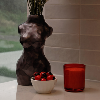 A decorative centerpiece featuring a black ceramic vase shaped like a woman’s torso. The vase sits on a counter next to a bowl of red cherries and a red candle.