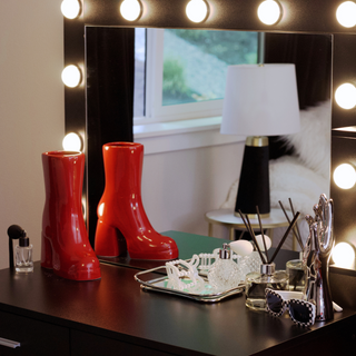 A vanity table with a rectangular mirror with bright lights. There is a a red boot vase sitting on the table next to the mirror.