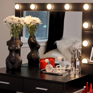 A lighted vanity table with a mirror. There are white flowers in a matte black body-shaped vase and perfume bottles on the vanity table.