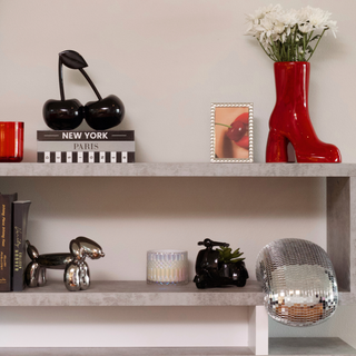 A shelf with decorative items. From right to left, there is a red boot vase holding white flowers, a framed photo of a woman holding a cherry, a silver disco ball, and three  books with a black cherry on top.