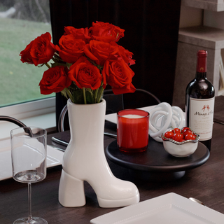 A white, ceramic boot vase filled with red flowers rests on a table. To the right of vase is a red candle and a bowl of cherries.