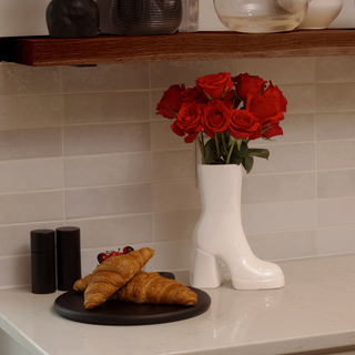 A close-up photo of a plate containing two flaky croissants. They are a light golden brown color and rest beside a white, ceramic boot vase. The vase holds a bunch of red flowers.