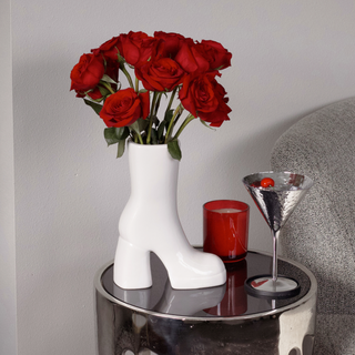 A white boot vase filled with red flowers sits on a table next to a martini glass and red candle. The table is positioned in front of a couch.