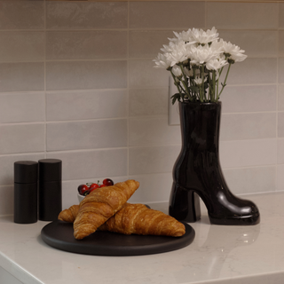 A close-up photo of a plate containing two flaky croissants. They are a light golden brown color and rest beside a black, ceramic boot vase. The vase holds a bunch of white flowers.