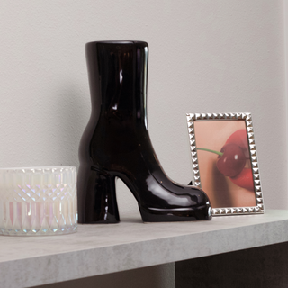 A black ceramic boot vase sits on a table next to a picture frame containing a photo of a woman with red lips.