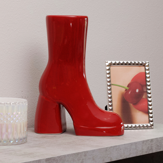 A red ceramic boot vase sits on a table next to a picture frame containing a photo of a woman with red lips. 
