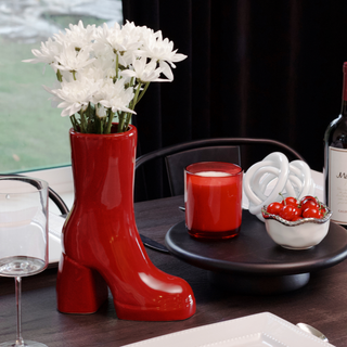 A red, ceramic boot vase filled with white flowers rests on a table. To the right of vase is a red candle and a bowl of cherries. 