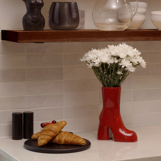 A close-up photo of a plate containing two flaky croissants. They are a light golden brown color and rest beside a red, ceramic boot vase. The vase holds a bunch of white flowers.